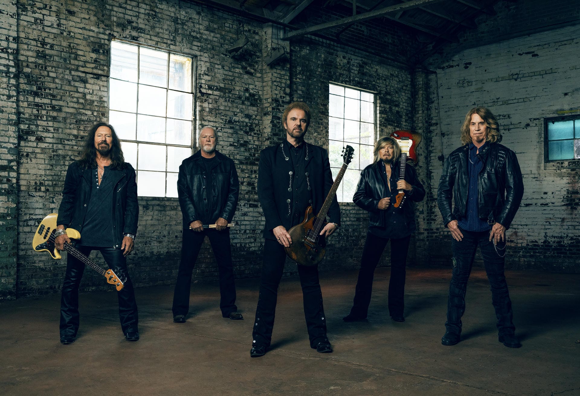 Five band members in black leather jackets posing in a brick-walled room.