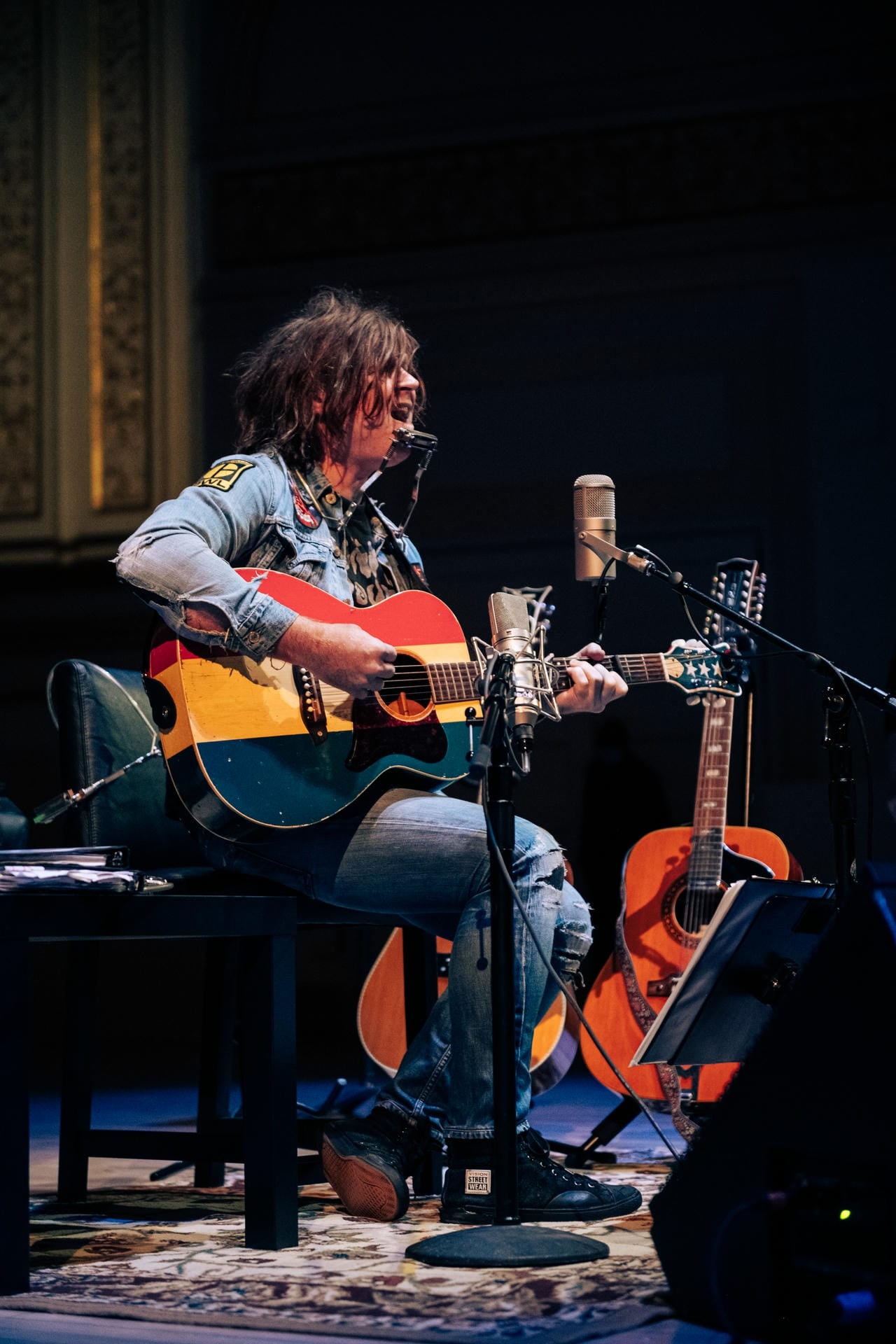 Person playing an acoustic guitar on a stage with a microphone and another guitar in the background.