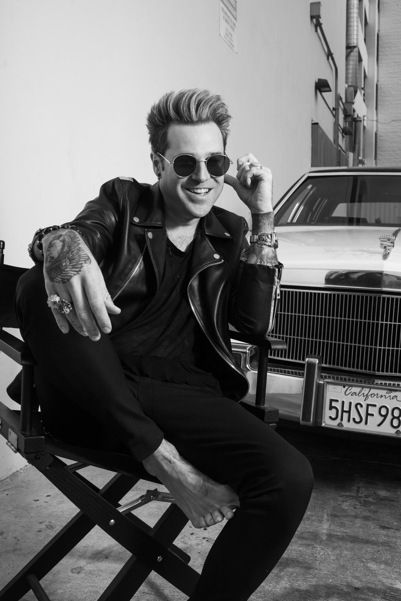 Man sitting on a chair in front of a vintage car, wearing sunglasses and a leather jacket.