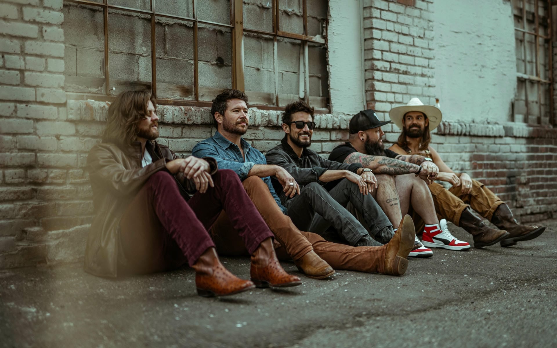 Group of six men sitting against a brick wall in an urban setting